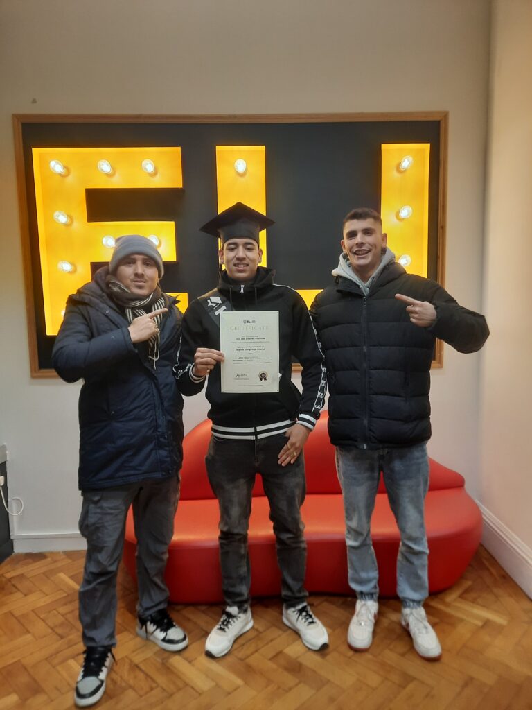 Tres personas posando en un aula frente a un letrero iluminado mientras uno de ellos sostiene un certificado y lleva birrete, mostrando un logro académico relacionado con estudiar inglés en el extranjero.