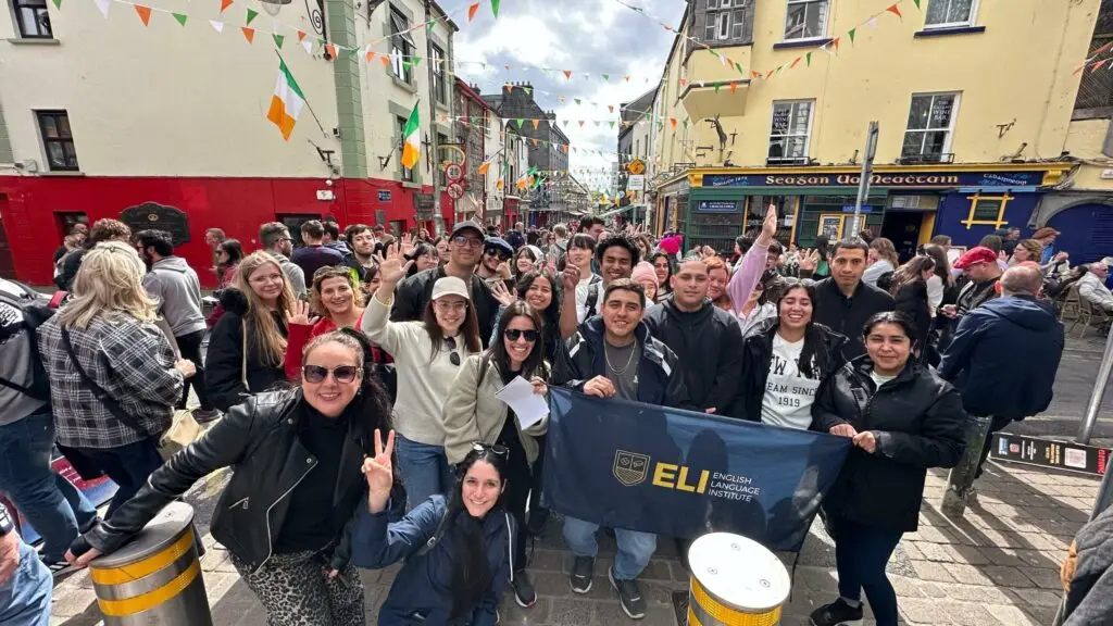 Gran grupo de estudiantes al aire libre en una calle decorada con banderines, sosteniendo una bandera del instituto y participando en una actividad cultural típica de quienes buscan estudiar inglés en el extranjero.