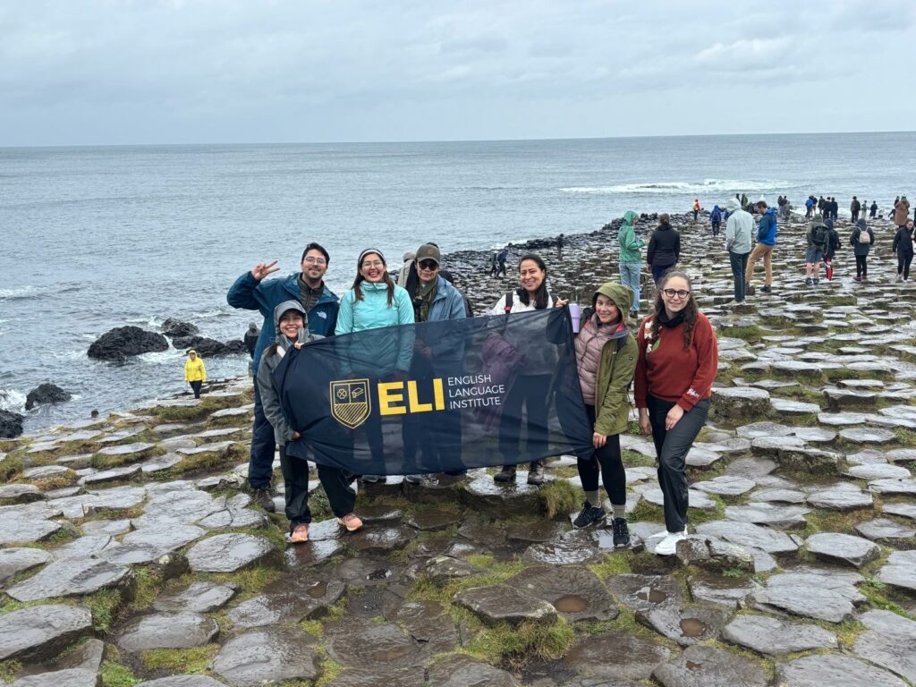 Alt: Grupo de visitantes posando sobre las columnas de basalto de la calzada del gigante (Giants Causeway), sosteniendo una bandera del English Language Institute frente al mar, con otros turistas caminando entre las formaciones rocosas al fondo.